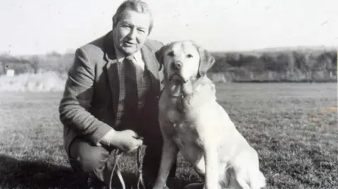 Guide Dogs A black and white image of a man kneeling in a field with a Labrador dog. The man is wearing a suit, checked shirt and striped tie. The dog is on a lead.