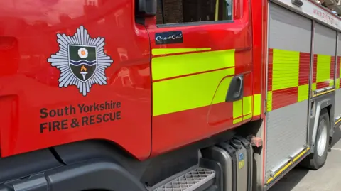 A South Yorkshire Fire and Rescue fire engine, with a fire service logo on the left of the picture. The vehicle is mainly red, with some fluorescent yellow squares.