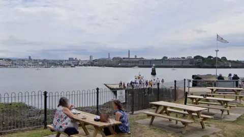 Google A street view of the Mount Edgcumbe slipway. There are two women sat at a picnic table on the grassy area and there are three other tables behind them. Below you can see people stood on the slipway and the top of the Cremyll ferry which is yellow. It is on the water, which is in the background along with other buildings on the other side of the sea.