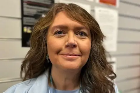 Janey has long, dark, wavy hair. She is wearing a pale blue suit jacket and dress and has an official NHS lanyard around her neck. She has tanned skin and a nose piercing. 