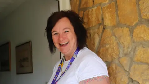 Caring Together Kerry Giles-Brown standing in the doorway of a home. She is wearing a white short-sleeved top with a purple lanyard ribbon around her neck. She is smiling and has short black hair parted over her forehead