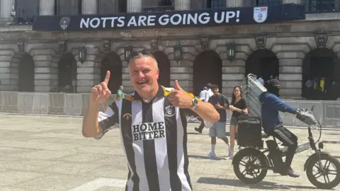 A man in a black and white-striped Notts County shirt in front of the council house with a banner that reads NOTTS ARE GOING UP