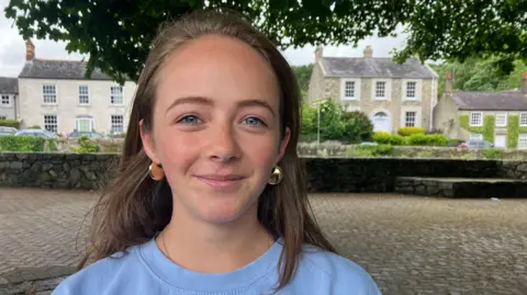 Orla Devlin, a young woman with blue eyes and long, fair hair, looking to the camera and smiling at the Fairy Glen in Rostrevor. She is wearing a light blue sweatshirt and gold earrings. A collection of Georgian houses line the river bank behind her.