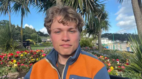 A man is sitting on a bench in a park, with flowers behind him and a blue sky. He is wearing an orange and blue top and he has light brown curly hair.