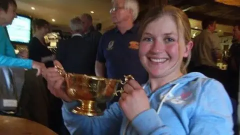 Jo Collinson A young woman wearing a light blue hoodie with blonde hair holds a gold cup as she smiles while sitting at a table in a busy Cotswolds pub