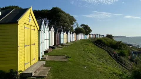 A row of colourful beach huts at the top of a grassy hill overlooking the sea. 