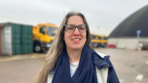 BBC/Becky Holmes Councillor Laura Robertson-Collins - she has green glasses and a blue and cream coat and long blonde hair. she is wearing gold earrings. you can see a line of gritters behind her. 