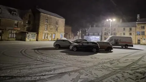 Image shows a high street with cars parked in the middle with snow on them.
