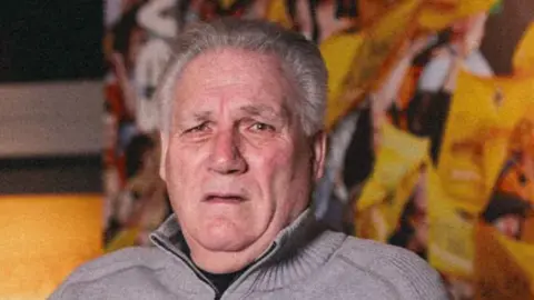 A man with thick grey hair is looking at the camera. He is wearing a wool-type cardigan with a collar, and is sitting in front of a yellow background.