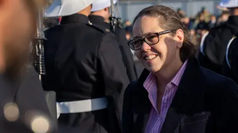 Sam des Forges has brown hair and glasses and is wearing a pink striped shirt. In the background is military personnel. 