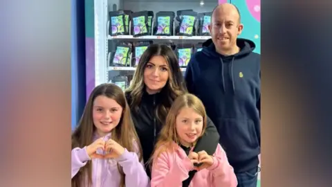 Little Dreamers A family photo of Amy and Derek and their two daughters standing in front of one of their vending machines. The girls are wearing light purple and pink hoodies and doing 'heart' hand gestures