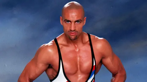 ITV/Shutterstock Daz Crawford in his days as a Gladiator. He's very muscular and wearing his white, black, blue and red singlet.