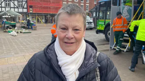 A woman with short hair in the centre of Lichfield. We can see bin workers at the mobile education centre behind her. She is smiling and wearing a white scarf and a black coat.