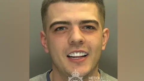 A custody photo of Coran Davies. Davies smiles looking at the camera. His white teeth are on show. 