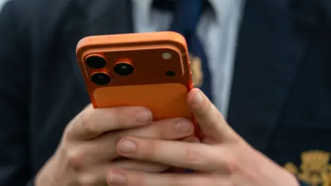 A close-up of a pupil's hands holding a metallic orange mobile phone. They are wearing a school blazer, shirt and tie.
