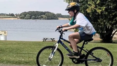 Danni Harper  Isaac smiling while riding his bike along a track that runs to the side of a lake. 