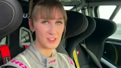 Lydia Walmsley sits in her race car dressed in grey race outfit and staring at the camera 