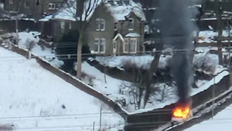 Brett Knowles A police van on fire on a snowy road, with the large house in the background.