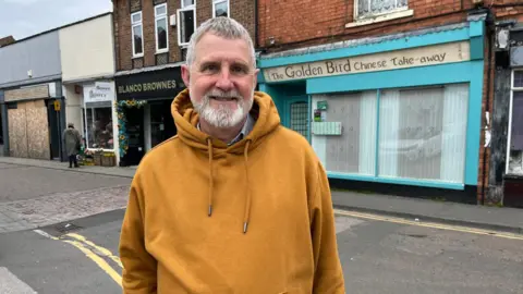 A man with grey hair and a beard wearing a yellow hoodie.