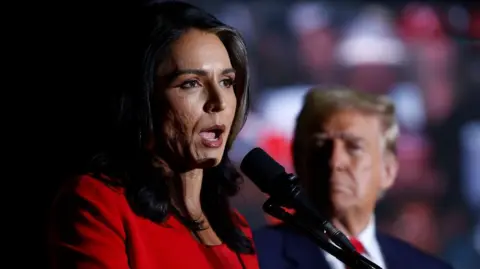 Getty Images Tulsi Gabbard, wearing a red pantsuit, speaks into a microphone. Trump's head is blurry in the background