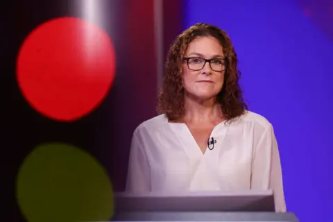 Bloomberg/Getty Images A woman with glasses and brown hair looks at the camera while wearing a white top, against a purple background.