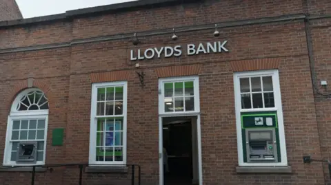 The photo shows a red-brick building. It has four Georgian-style windows and above them are the words 'LLOYDS BANK'. To the right is a cash machine, with the Lloyds logo on it. There is also a cash machine to the left.