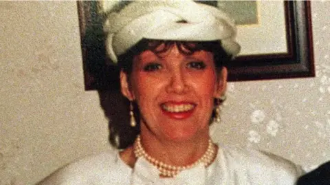 Family Handout A woman smiling at the camera. She is wearing a white dress and hat. She is wearing pearl earrings and necklace. She is standing in front of a framed picture hanging on a wall.