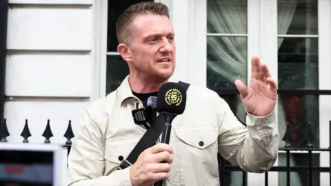 Tommy Robinson, standing outside Westminster Magistrates' Court, in London, talking into a black microphone, with a golden lion head on it, with his left hand raised. He is wearing a light jacket, a black T-shirt and has short dark hair and short beard. He is standing by black railings. 
