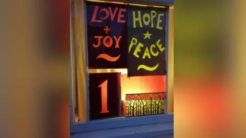 DRARA A house window with a decoration of a the words "Love + Joy" and "Hope and Peace" on display. The number 1 is displayed beside it.