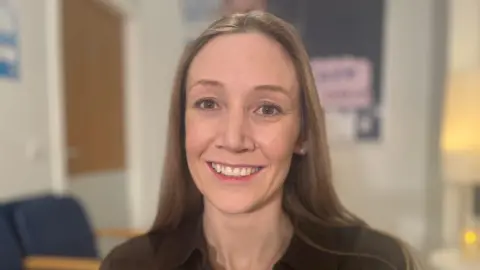 Smiling Laura Corcoran has long brown hair. She is sitting in a hospital or doctor's waiting room