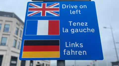A blue sign with the British, French and German flags and the message 'drive on the left' in English, French and German.