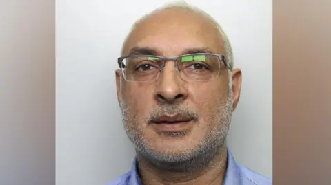 A man in his 50s, pictured in a police mugshot. He has very thin white hair, white stubble and glasses. He is wearing a blue buttoned shirt and staring directly at the camera.