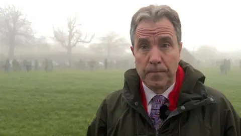 Dan Norris is on the right of the picture looking at the camera. He is wearing a green coat, with a shirt and tie underneath. Behind him on the field are small groups of people and a layer of fog.