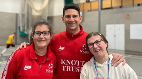 Mark and two female players stand together inside an indoor sports hall. The girls are wearing red sports jackets with “Knott Trust” branding, while Mark is wearing a light-coloured hoodie with a lanyard. In the background, badminton players are visible on the court, and spectators are watching from an upper gallery.