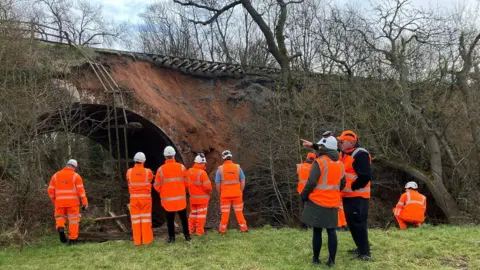 Eight people are on some grass looking up at a landslip on top of a railway bridge. There is fencing hanging off the top of the bridge and earth has fallen away. They are taking pictures and pointing up at the tracks which are suspended over the edge of the bridge