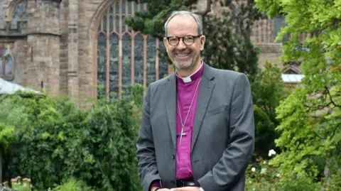 Diocese of Hereford The Bishop of Hereford, a man with short grey hair and glasses, stands in front of what looks like a cathedral building. He is wearing a grey suit jacket over a purple priest's smock with a white collar and has a cross around his neck. He is smiling at the camera.