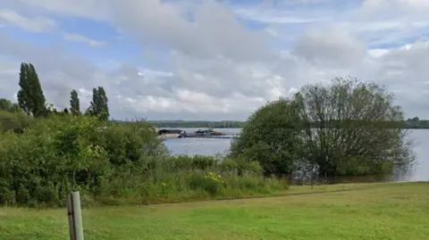 A reservoir is seen behind a lawned area with trees and other shrubbery. On the water is a pontoon and a boathouse.