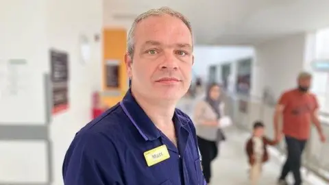 A middle-aged man with short grey hair stands in a white hospital corridor. He is wearing a navy polo shirt and a yellow name badge which reads "Matt". A couple and their small child can be seen in the blurred background behind him.