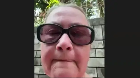 BBC Angela has her hair tied back and is wearing sunglasses. There is a brick wall behind her.