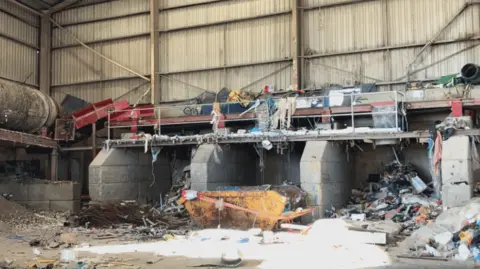 Lara King A large warehouse,. with corrugated metal walls and concrete fixtures. There are piles of scrap and a sorting line.