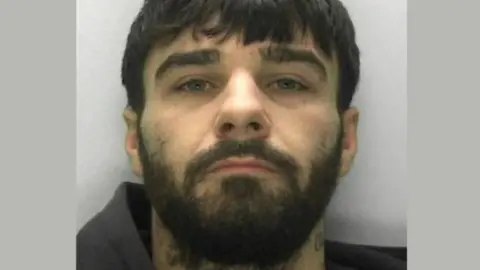 Connor Parsons looks into the camera for his police custody image. He has black hair, a thick beard and wears what looks like a black hoodie.