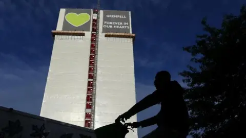 Reuters Grenfell Tower after the fire and wrapped in plastic, with the silhouette of a man riding a bike in front