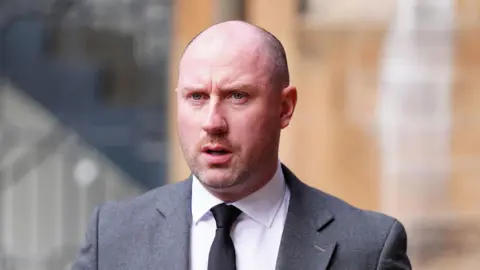 PA Media Neil Gray is bald and wears a grey suit with a white shirt and black tie. He looks ahead while walking outside a building
