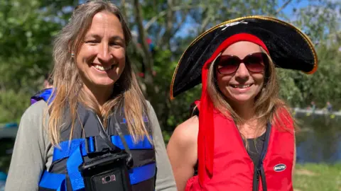 Jenny Kirk/BBC Jacky and Mel smile at the camera with the river behind them. Jacky has long fair straight hair and wears a blue life jacket with bright blue straps, over a grey hoody. Mel wears a black vest with a bright red life jacket over it, sunglasses, and a wide brimmed pirate hat.