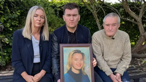 Joseph Brett wearing black and holding a framed photograph of his sister, who has blonde hair. Either side of Mr Brett are Noel Brett, wearing a beige jumper, and Pauline Nimmo, wearing a blazer and pale blue top. They are all sitting on a bench and look serious.