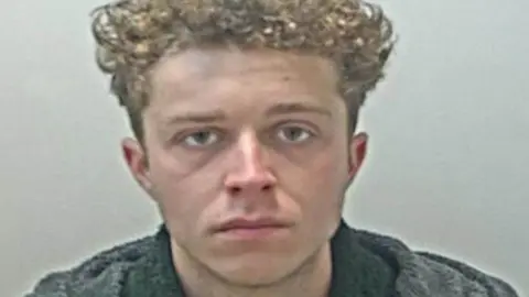 Lancashire Police Police custody image of Frankie Bromley, who has light brown curly hair nd is wearing a black jumper and grey coat.