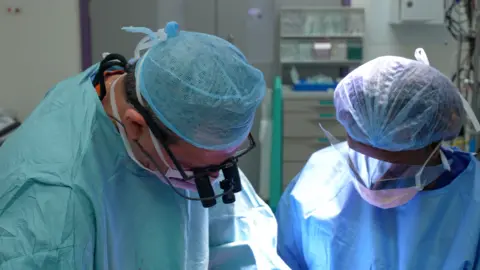 BBC News Two members of the surgical team at the Royal Papworth Hospital perform a double lung transplantation. The clinicians, a man and a woman, are wearing mint green and blue scrubs and hair nets. In the background we see elements of the operating theatre out of focus