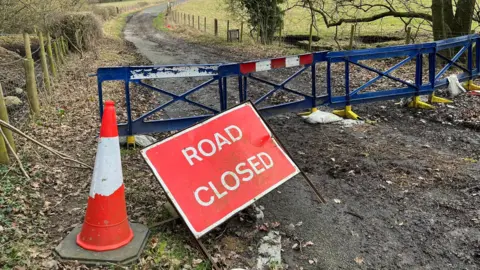 A road closure with blue barriers spanning a road and a red 'Road Closed' sign which has a slight green sheen of moss. 