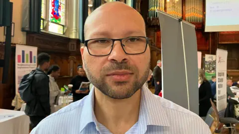 Sameh Hassan, with short hair and black glasses in a blue striped shirt. Behind him is a crowded room in the Guildhall in Londonderry.