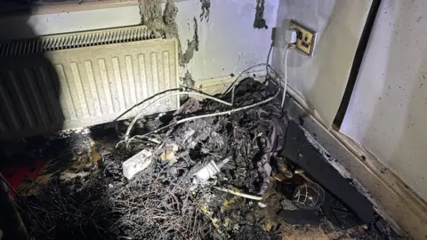 A bundle of wires are on the floor and are burnt from a fire. There is a plug socket on the right had side and a white radiator on the left. 